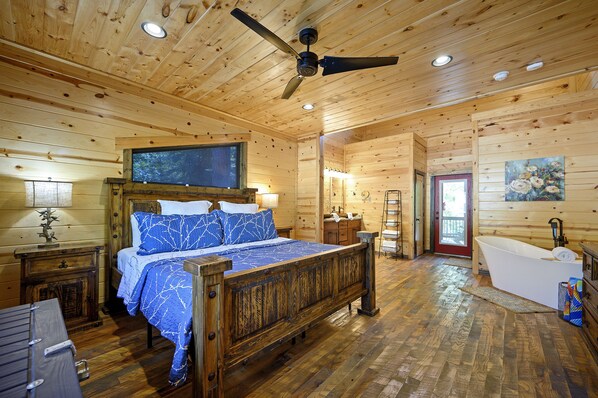 Une chambre dans une cabane en bois, comprenant un lit, un ventilateur de plafond, une commode en bois, une table de chevet, une fenêtre et une porte.