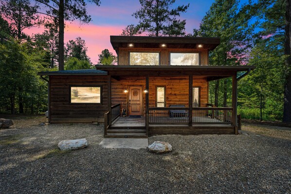 A two-story log cabin with a porch, large windows, and a gravel driveway. - Waymaker, Couples, Hot Tub, Firepit, Soaker Tub (Broken Bow)