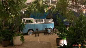 Un fourgon bleu est garé dans une serre, où se trouvent des plantes en pot et des tabourets.
