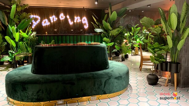 Un espace détente aux couleurs vertes, agrémenté d’un néon lumineux indiquant le mot « Dancing », dans une pièce ornée de plantes en pot et dotée d’un sol carrelé.