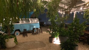 Un fourgon bleu est garé dans une zone sablonneuse, à proximité de plantes en pot et de tabourets en bois.