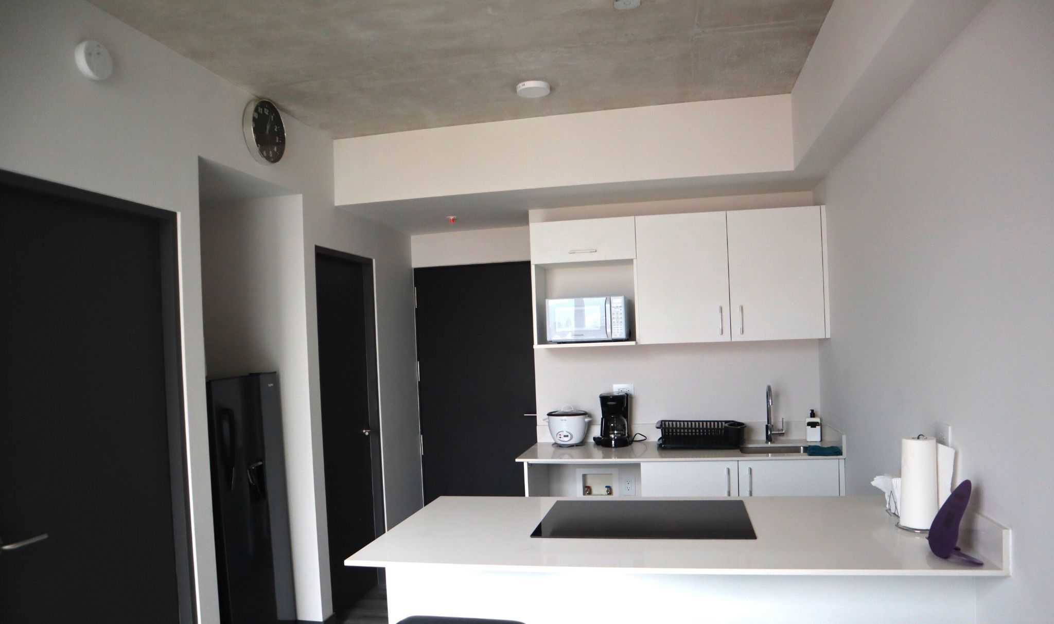 A modern kitchen with white cabinetry, a built-in microwave, a coffee maker, and a sink. There are two black bar stools at the counter.