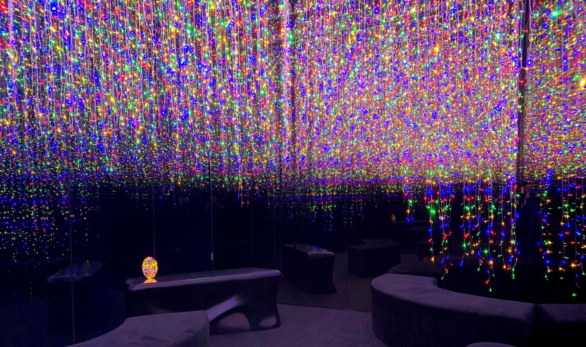 Una habitación con un techo cubierto de luces multicolores, una pequeña mesa con un adorno en forma de huevo y varias áreas de descanso en tonos oscuros.