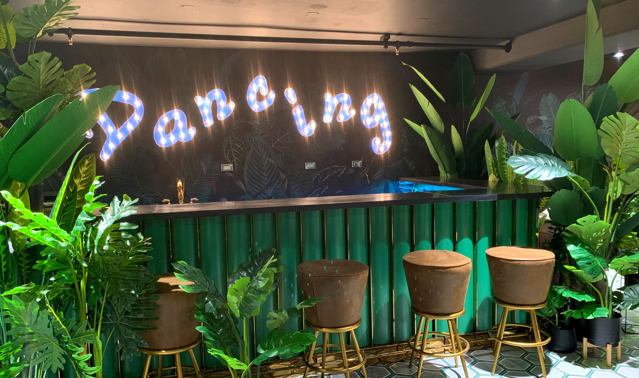 A bar area with a neon sign reading "Panning," surrounded by lush green plants and three stools.