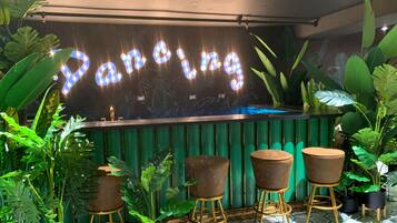 A bar area with a neon sign reading "Panning," surrounded by lush green plants and three stools.