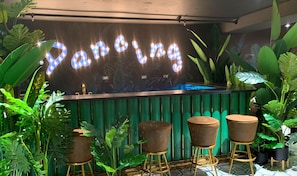 A bar area with a neon sign reading "Panning," surrounded by lush green plants and three stools. - Experiencia Mágica al Mejor Precio (San José)