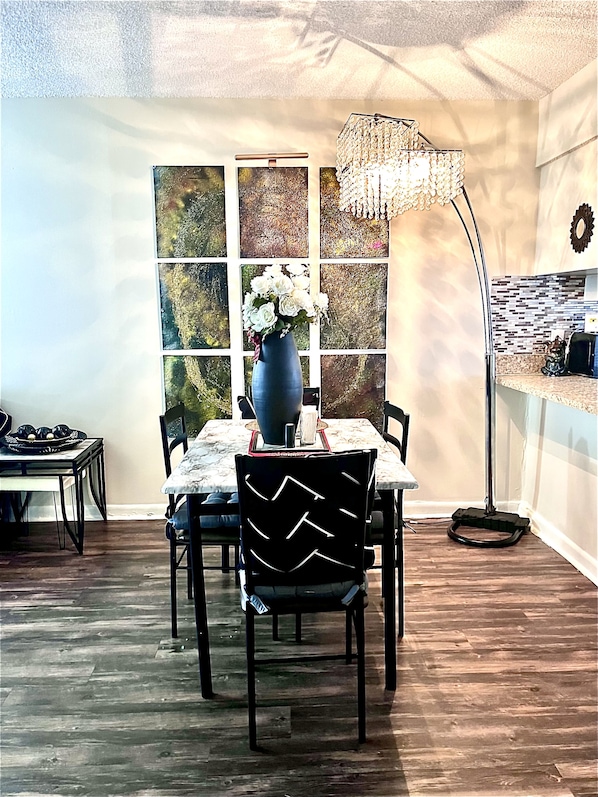 Un comedor con una mesa dispuesta para cuatro personas, rodeada de sillas con estampado blanco y negro. Un jarrón con flores blancas está sobre la mesa, y una lámpara de pie se encuentra a la derecha.