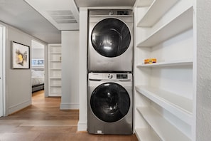 A laundry room with two stacked front-loading washing machines, a shelf with a detergent box, and a hallway leading to a bedroom.