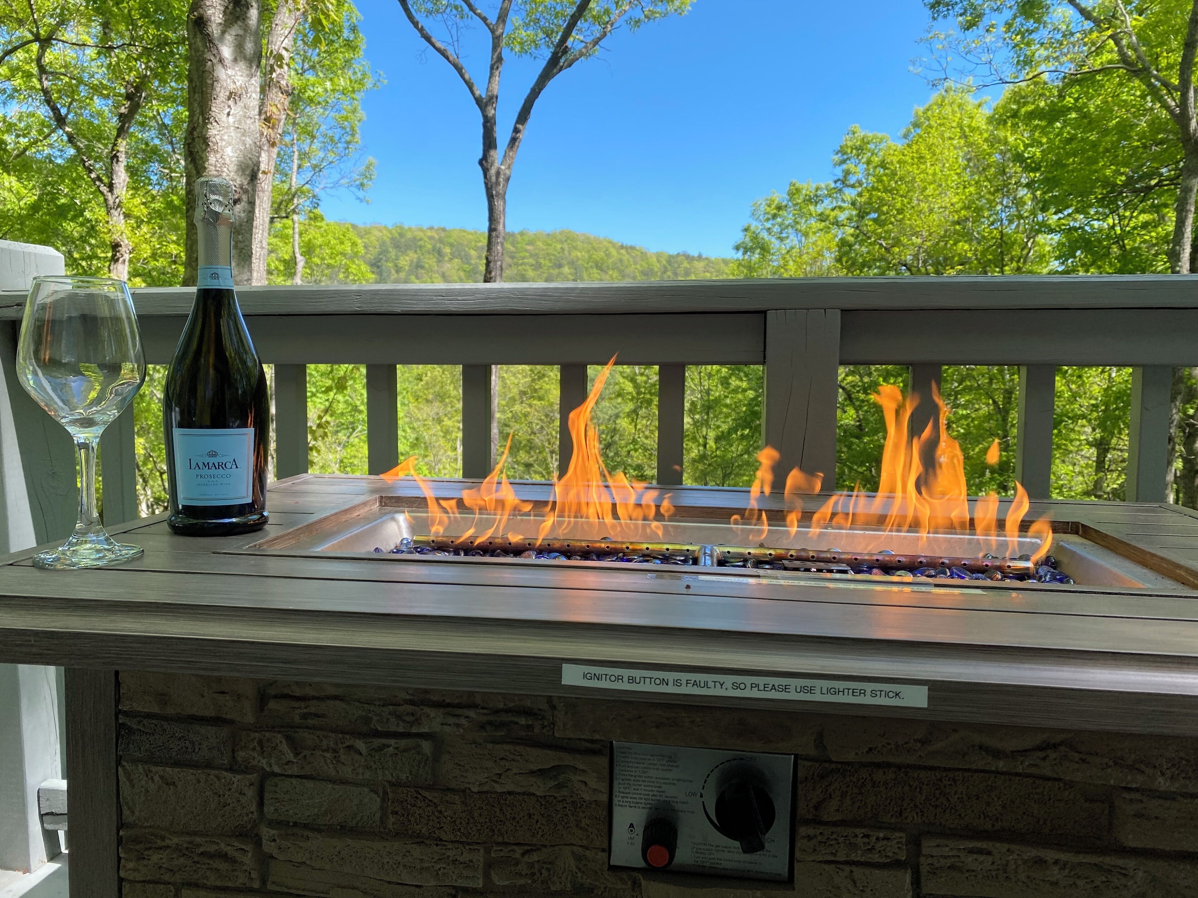 Una parrilla con llamas, una botella de vino y una copa sobre una terraza de madera, con vista a árboles y colinas.