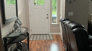 Un couloir doté d’un plancher en bois, d’une porte blanche, d’une table à plateau en verre ornée d’objets décoratifs, d’un canapé en cuir noir et d’un luminaire fixé au mur.