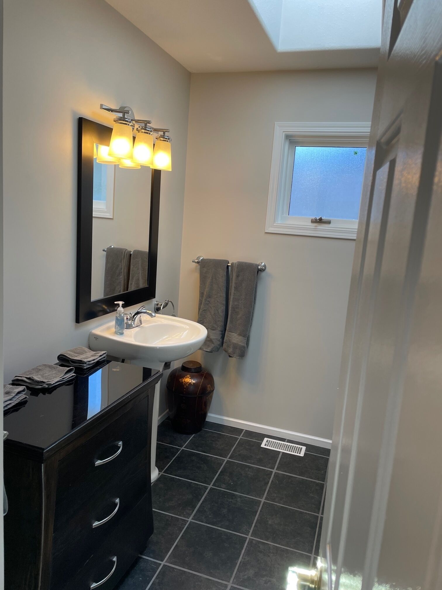 A modern bathroom with a white sink, dark tiles, a large mirror with lights, and a window.