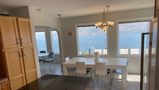 Una cocina con encimera negra, armarios de madera, un comedor con sillas blancas y grandes ventanas con vistas al agua.