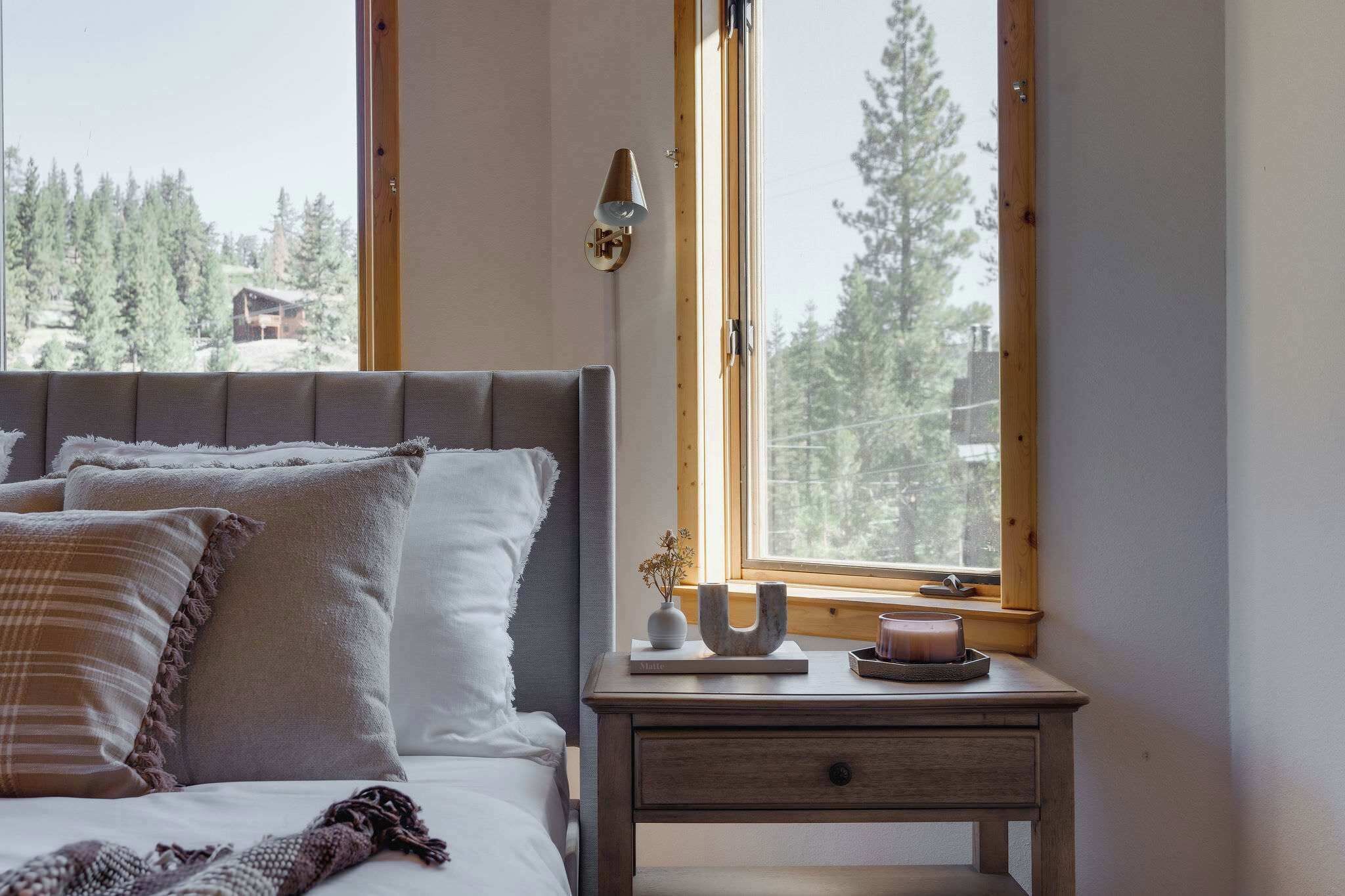 A bedroom with a bed, bedside table, and a window with a view of trees and a cabin.