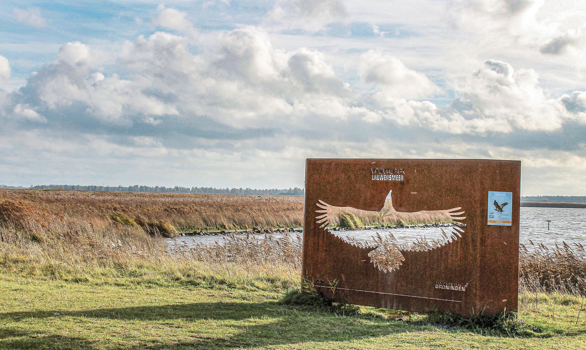 Een verroest metalen bord met een vogeldecoratie en tekst, gelegen in een grasrijk gebied in de buurt van water.