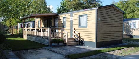 A single-story mobile home with a porch and a small deck, surrounded by a grassy yard and trees.