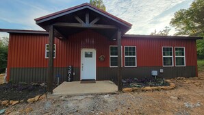 A red and brown metal building with a white door, windows, and a porch. - Cozy cabin 16 miles from Razorback Stadium great for game weekends. No pets. (Elkins)