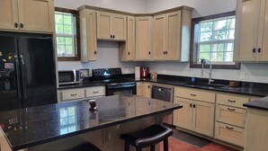 A kitchen with light wooden cabinets, a black refrigerator, a stove, and a sink. There are two bar stools and a window providing natural light. - Cozy cabin 16 miles from Razorback Stadium great for game weekends. No pets. (Elkins)