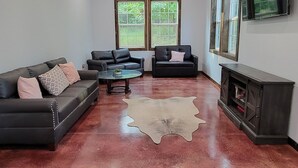 A spacious living room with a polished red floor, two black leather sofas, a coffee table, and a ceiling fan. - Cozy cabin 16 miles from Razorback Stadium great for game weekends. No pets. (Elkins)
