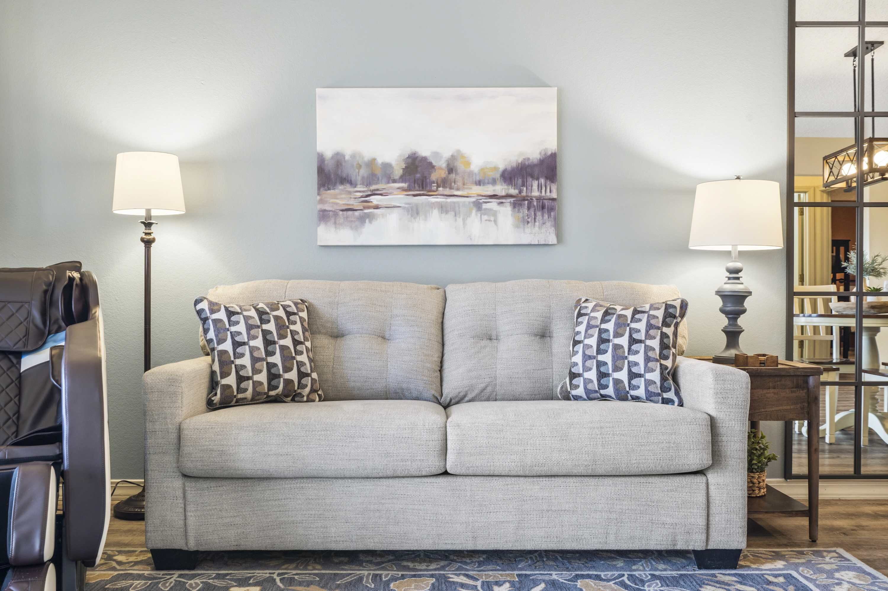 A living room with a grey sofa, patterned throw pillows, a painting above, and two floor lamps.