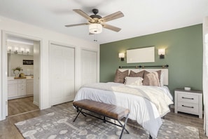 Quarto com cama, um banquinho, mesa de cabeceira, ventilador de teto e espelho.