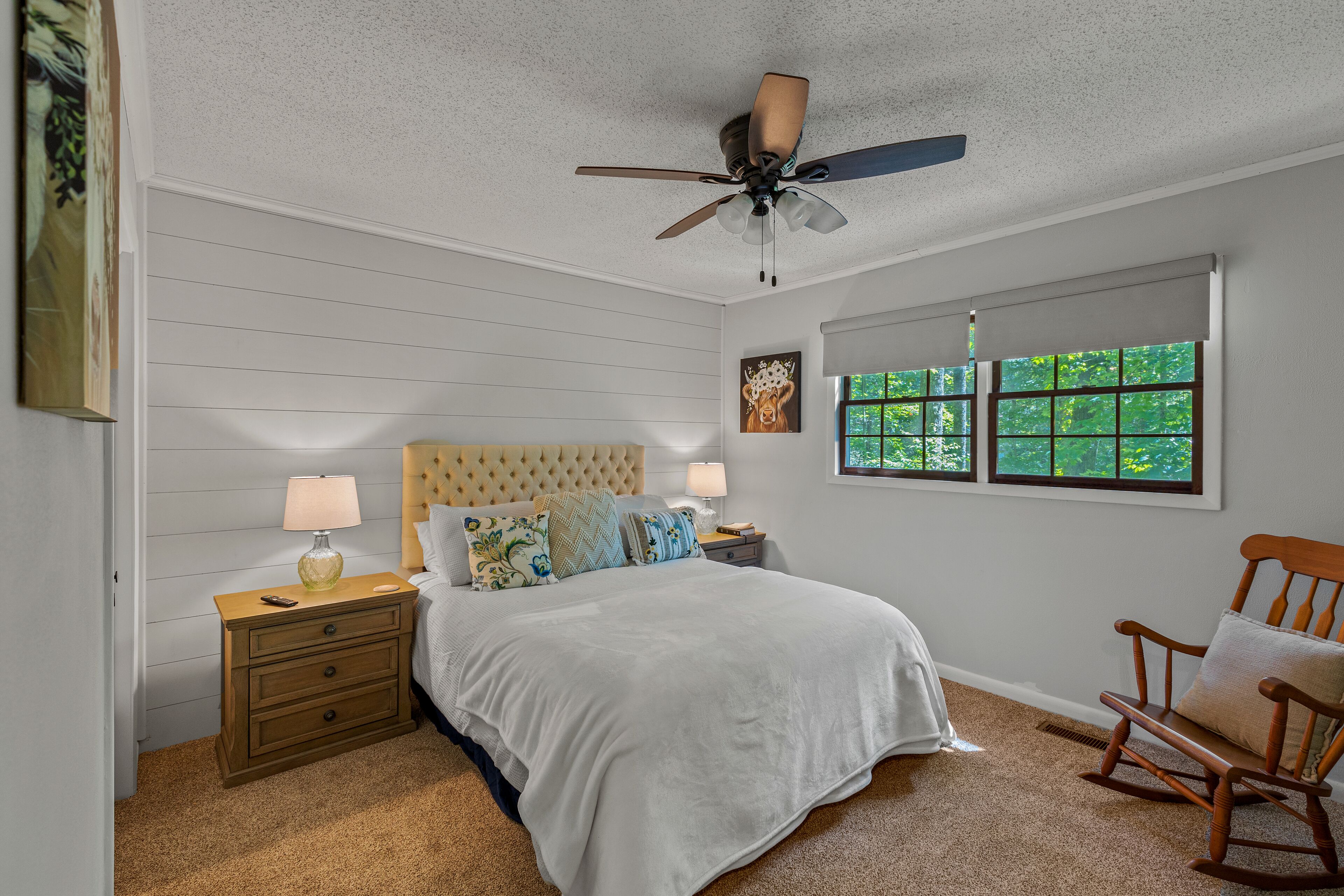 Une chambre à coucher comprenant un lit, des tables de chevet, un fauteuil à bascule, un ventilateur de plafond et une fenêtre donnant sur un paysage verdoyant.