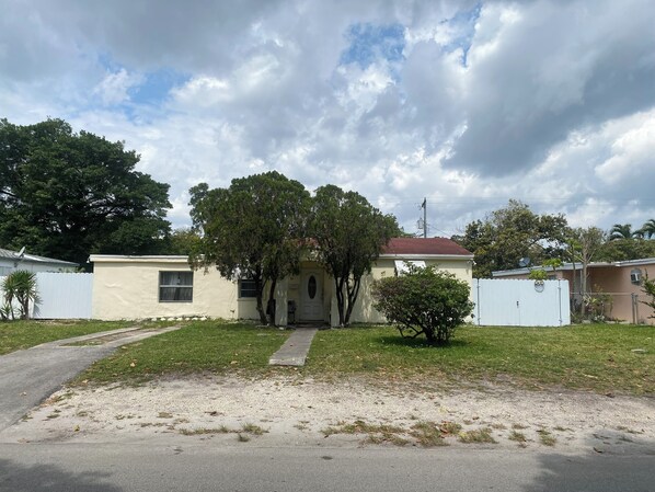 A single-story house with a red roof, a front yard with grass and a pathway leading to the door, and a white fence in the background. - Tiny homes Miami (North Miami)