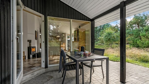 An outdoor dining area with a table and chairs, a glass door leading to a room with a fireplace, and a view of the surrounding greenery.