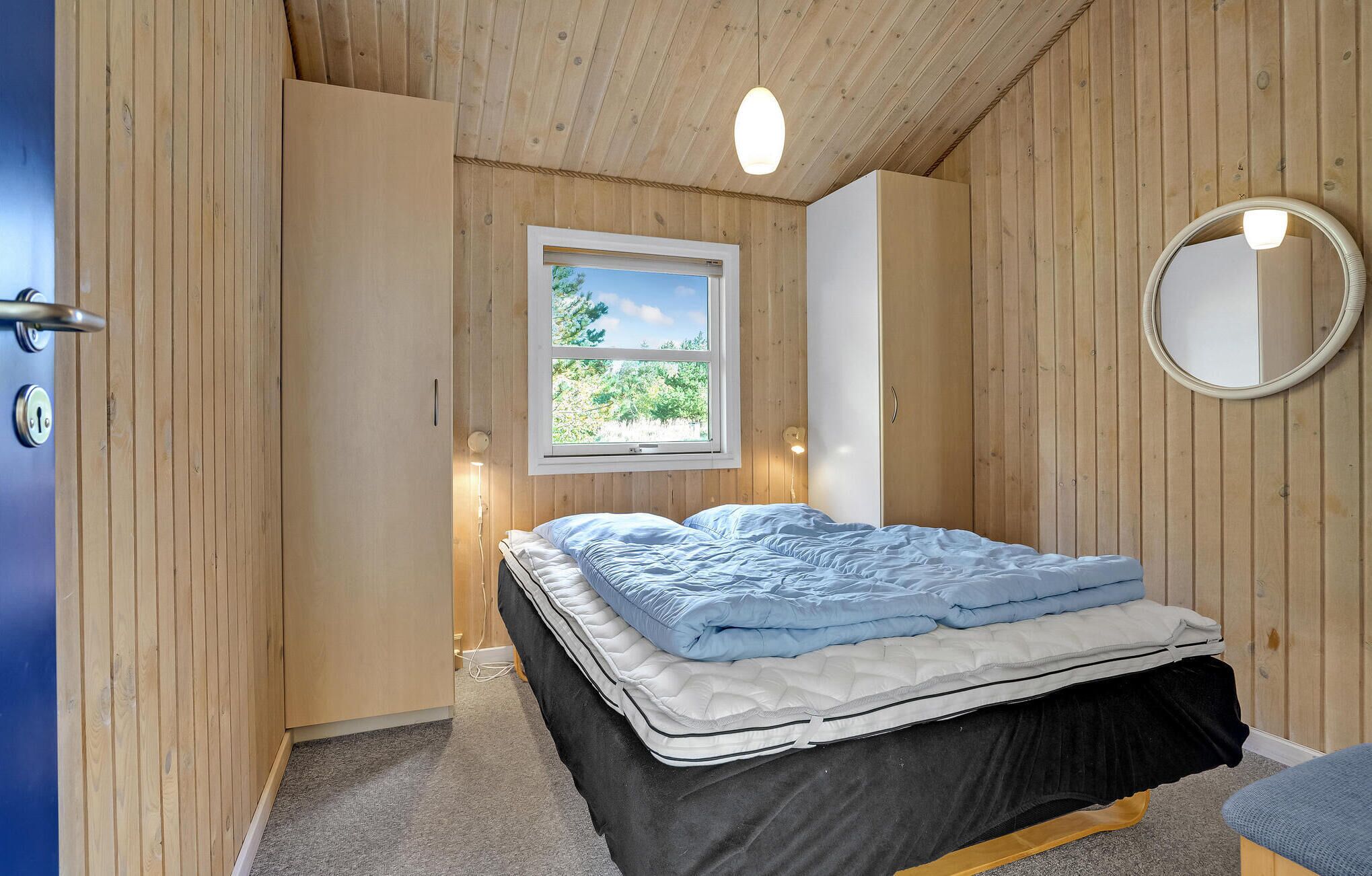 Quarto com cama, janela com vista, espelho redondo e paredes de madeira.