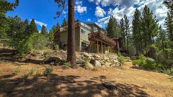 A two-story cabin with a wooden deck and stone foundation, surrounded by a forested area.