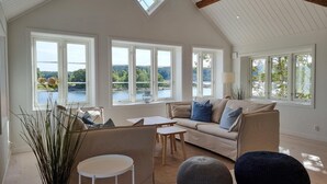 A spacious living room with a high ceiling, large windows offering a view of a lake and hills, a beige sofa with blue cushions, a small round table, and a tall plant.