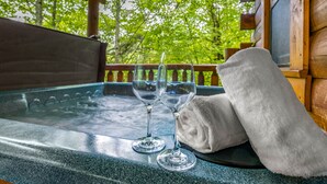 Een houten chalet met een hottub en twee lege wijnglazen op een tafel, omgeven door weelderige groene bomen.