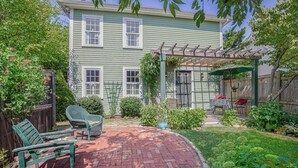 A two-story house with a brick patio, green outdoor furniture, and a pergola with a green umbrella.