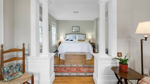 A bedroom with a bed, nightstands, a lamp, a chair, a table, and a potted plant.