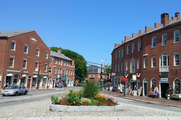 A historic street with brick buildings, a cobblestone street, and a flower bed in the center. - Secret Garden (Newburyport)
