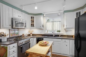 A kitchen with white cabinets, a stainless steel oven, a black refrigerator, and a wooden table with a fruit bowl. - Secret Garden (Newburyport)