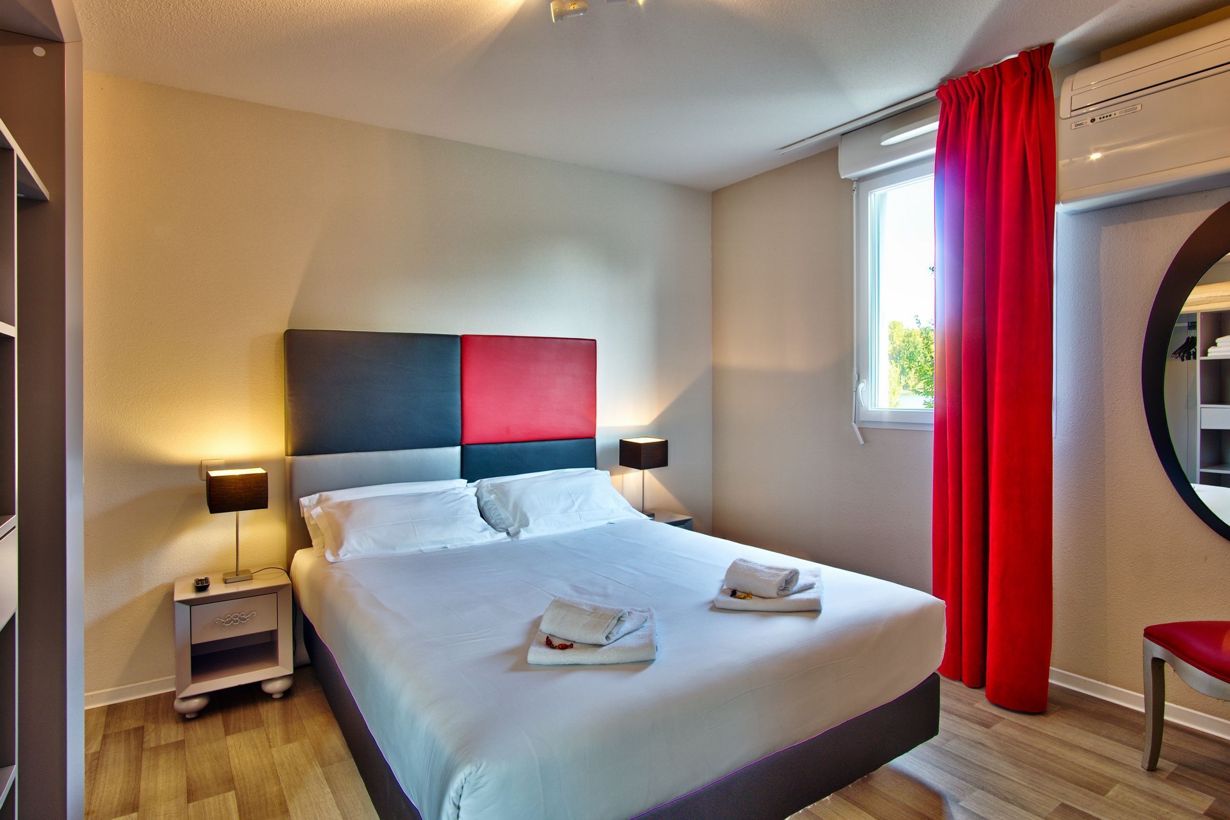 A modern hotel room with a large bed, two bedside tables with lamps, a red curtain, and a round mirror.