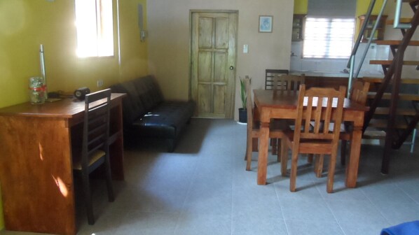 A living room with a sofa, wooden dining table, and chairs. There is a door in the background and a window with blinds.