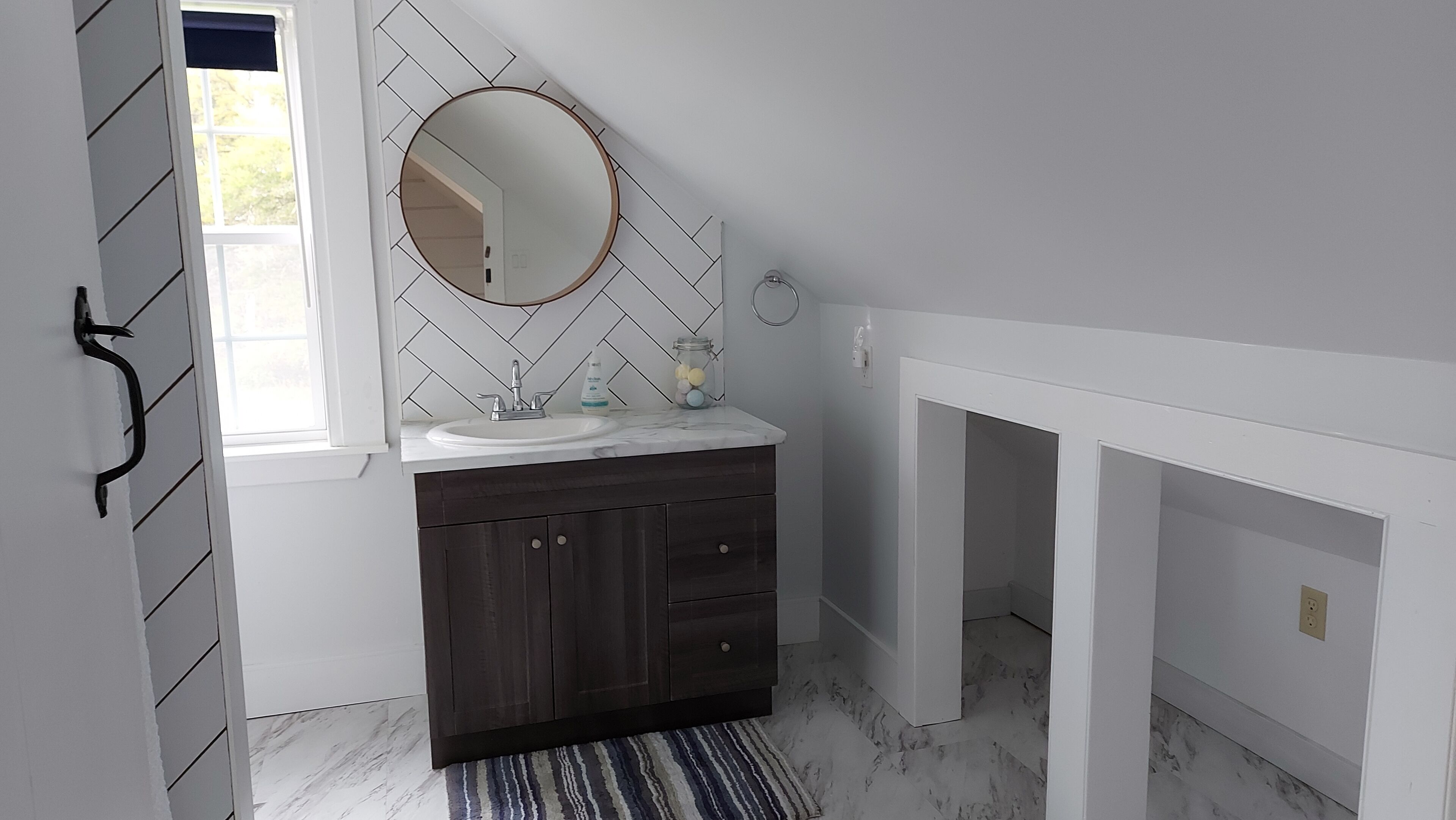 Un baño con un salpicadero de azulejos blancos tipo espiga, un espejo redondo, un mueble de madera oscura y una alfombra a rayas.
