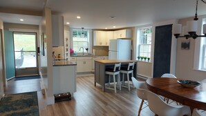 A modern kitchen with white cabinets, a central island, and a dining area with a wooden table and white chairs. - Cozy Harbour House - Scenic-relaxing-waterfront (Municipality of the District of Guysborough)