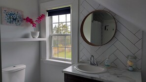Une salle de bain avec des toilettes blanches, une fenêtre donnant sur des arbres, un lavabo avec un miroir et une plante en pot aux fleurs roses.