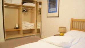 Quarto com uma cama, uma mesinha de cabeceira de madeira com abajur, e um guarda-roupa aberto com roupas penduradas e dobradas.