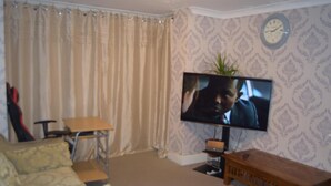 Sala de estar com televisão, um banco de madeira, uma mesa pequena e parede estampada.