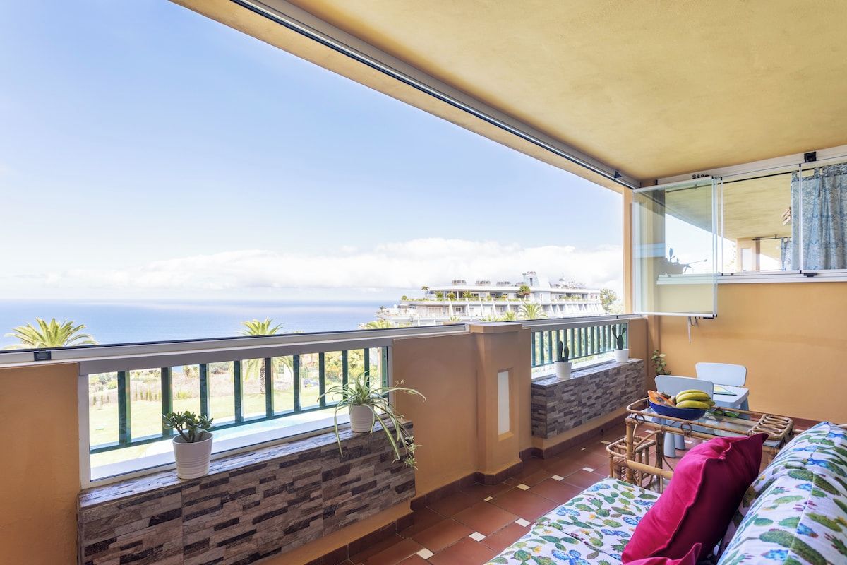 A balcony with a view of the sea, a sofa, a table with a basket, and potted plants.