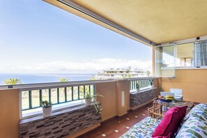 A balcony with a view of the sea, a sofa, a table with a basket, and potted plants.