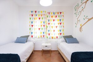Une chambre avec deux lits, une table de chevet, une fenêtre ornée de rideaux triangulaires aux couleurs vives et une fresque murale représentant un arbre.