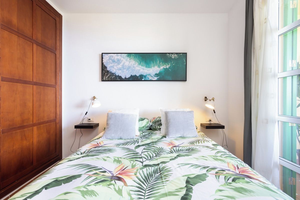 A bedroom with a bed, a wooden wardrobe, a wall-mounted picture, bedside lamps, and a window with curtains.