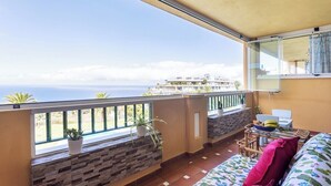Balcón con vista al mar, un sofá, una mesa con una computadora portátil y plantas en macetas.
