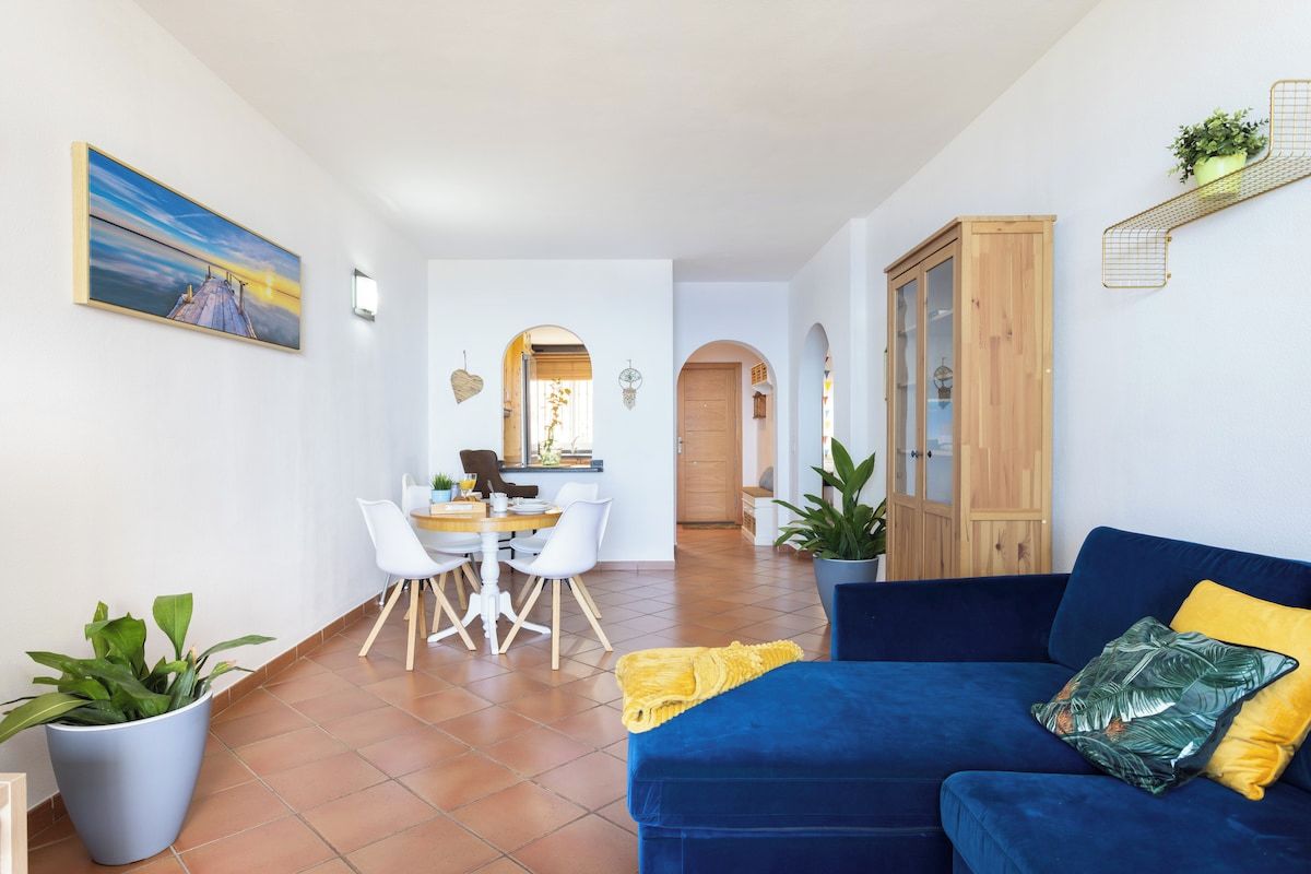 A living room with a blue sofa, a dining area with a table and chairs, and a kitchen in the background.
