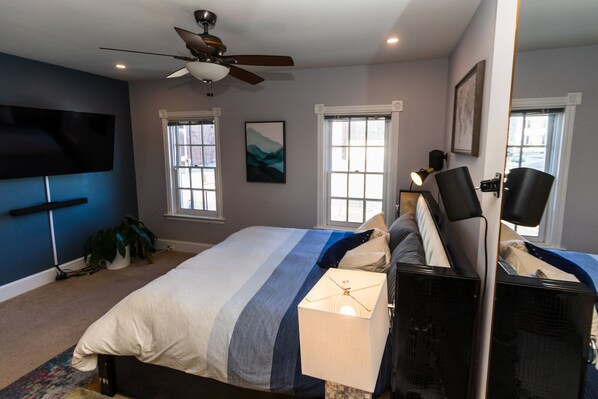 Un dormitorio con cama, ventilador de techo, televisor de pantalla plana y dos ventanas.