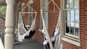 Une véranda avec deux hamacs en macramé suspendus au plafond, un bâtiment en briques et une balustrade blanche.